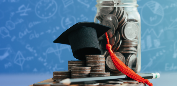 Stack of quarters wears a graduation cap next to a jar of coins in front of classroom blackboard