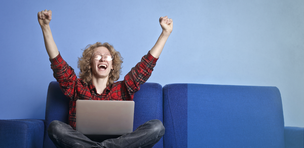 Young man wearing glasses and red print shirt with both arms up excitedly with laptop in lap while seated on a sofa