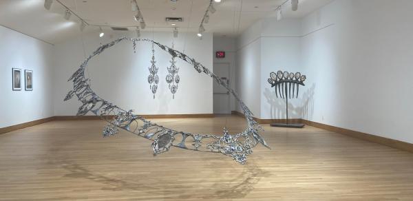 Gallery installation shot showing a colossal hanging metal sculptures in the shape of a necklace, earrings, and a comb.