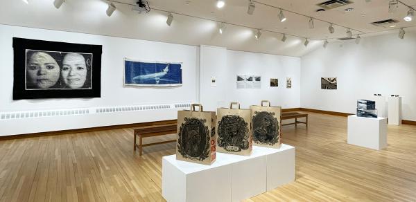 view of artwork installed inside gallery: pieces are installed on the walls; on the far left is a piece containing two faces, the piece next to it is an image of a plane. In the center of the gallery are three oversized brown grocery bags with images of field workers.