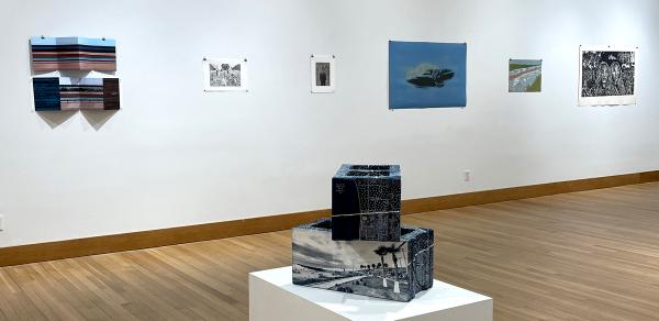 view of artwork installed inside gallery: Six works on paper are installed in the wall of the gallery. in the middle of the image are two paper sculptures in the form of cinderblocks one on top of the other and both with images of the US-Mexico border in San Diego, CA.