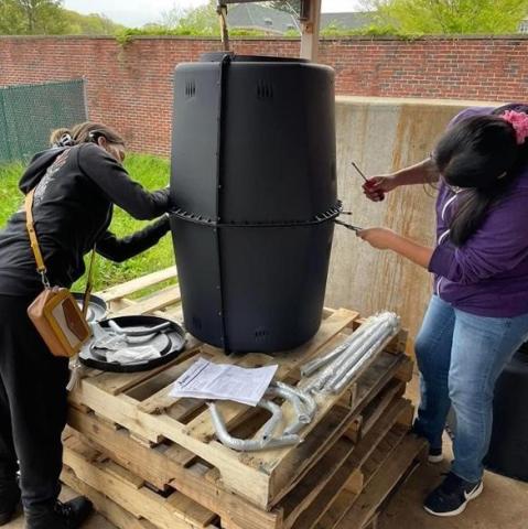 2 students are building a compost bin . They are each using a screwdriver to attach the two sections of the barrels together.